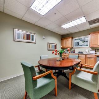 A small office meeting room with a round wooden table and several chairs, featuring artwork on the walls and a kitchenette in the background.