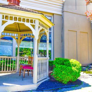 A charming gazebo with a wooden chair is surrounded by lush greenery on a sunny day.