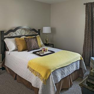 A cozy bedroom with a neatly made bed featuring decorative pillows and a yellow blanket, accompanied by a nightstand with a lamp and tray.