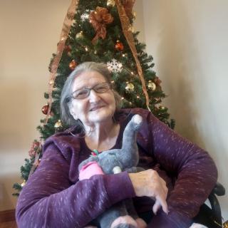 An elderly woman sitting in front of a Christmas tree, holding a stuffed animal.