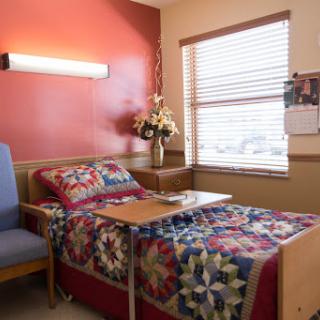 A cozy hospital patient room featuring a bed with a quilt, a chair, and floral decorations by the window.