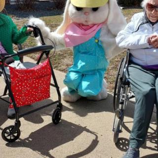 An Easter bunny mascot poses with two elderly individuals, one using a walker and the other in a wheelchair, outside on a sunny day.