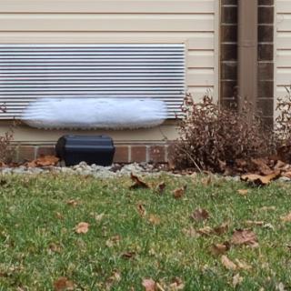 A vent with snow accumulation below it, surrounded by fallen leaves and grass.
