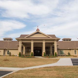 A large building with a grand entrance featuring columns and landscaped surroundings under a partly cloudy sky.