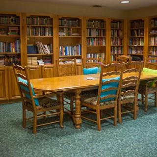 A cozy library room with a wooden table surrounded by chairs, and bookshelves filled with books.