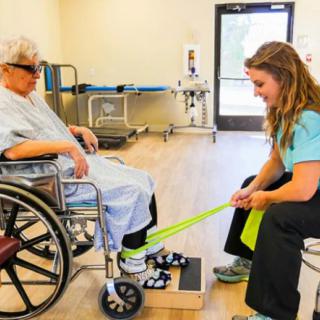 A healthcare professional assists a patient in a wheelchair with physical therapy exercises using an exercise band.