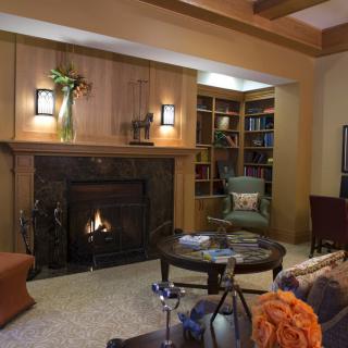 A cozy living room with a warm fireplace, surrounded by wood paneling and stylish furniture, featuring bookshelves and decorative elements.