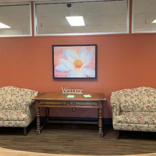 A cozy waiting area featuring floral artwork against an orange wall, complemented by two upholstered chairs and a wooden table.