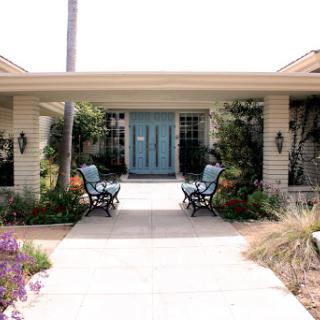 A charming house entrance with a blue door, surrounded by lush landscaping and comfortable outdoor seating.