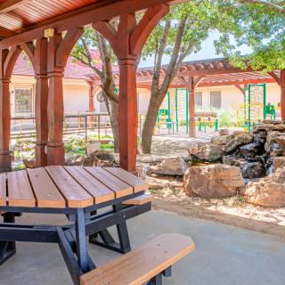 A serene outdoor setting featuring a wooden gazebo with a picnic table, surrounded by trees and rocks.