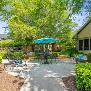 A serene outdoor patio area with chairs and a table under an umbrella, surrounded by lush greenery.