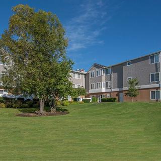A residential apartment building with a well-maintained lawn and surrounding trees under a clear blue sky.