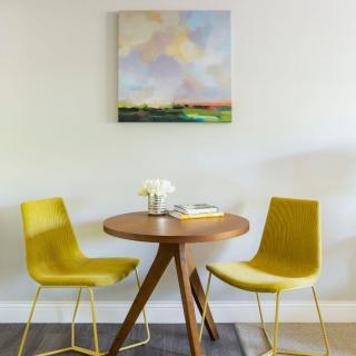 A modern dining area featuring two yellow chairs and a round wooden table, complemented by a colorful painting on the wall.