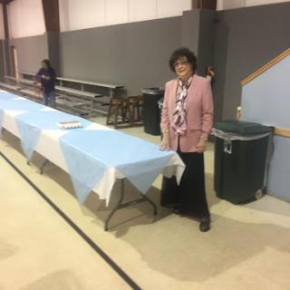 A woman in a pink jacket stands next to an empty table covered with a blue tablecloth in an indoor setting.