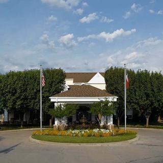 A building entrance with a circular driveway, landscaped garden, and flags under a cloudy sky.
