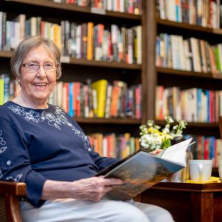 An elderly woman sits comfortably in a library, enjoying a book with a cup of tea and flowers nearby.