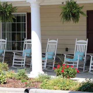 A row of white rocking chairs on a porch adorned with hanging plants and flowers.