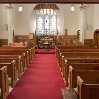 Interior view of a church with wooden pews, red carpet, and a stained glass window behind the altar.