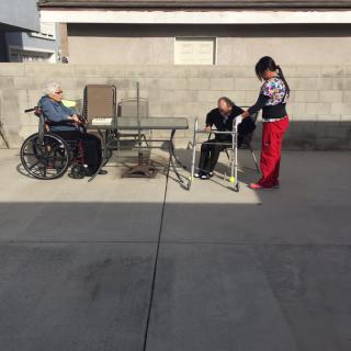 Two elderly individuals are sitting outdoors, one in a wheelchair and the other with a walker, accompanied by a caregiver on a sunny concrete patio.