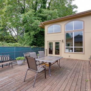 A spacious outdoor patio with a wooden deck, featuring a table and chairs, adjacent to a modern house surrounded by greenery.