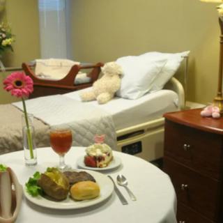 A hospital room with a bed, teddy bear, and a table set with a meal and flowers.
