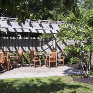 A shaded patio area with wooden chairs and a table under a pergola, surrounded by greenery.