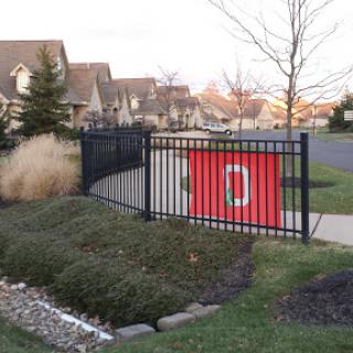A decorative red holiday banner hangs on a black fence in a residential neighborhood, surrounded by a well-manicured lawn.