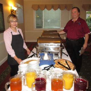 Two people standing by a buffet table with various drinks and covered dishes ready for serving.
