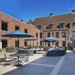 A sunny courtyard featuring brick buildings, outdoor seating with umbrellas, and clear blue skies.