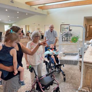 A family gathers indoors, with seniors in wheelchairs and walkers interacting with children.