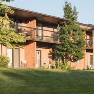 A two-story apartment building with brick exterior and balconies, surrounded by a well-kept lawn and trees.