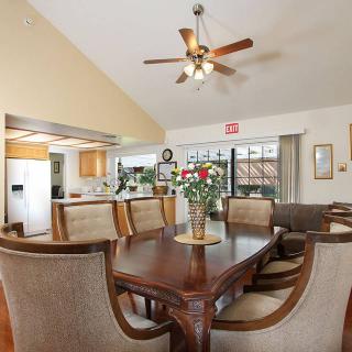 A spacious dining room with a wooden table, upholstered chairs, and a ceiling fan, adjacent to a kitchen.