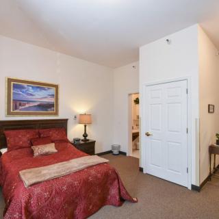 A cozy bedroom featuring a red bedspread, warm lighting from a table lamp, and wall art above the bed.