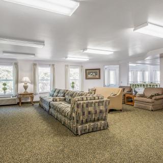 A spacious living room with multiple sofas, a carpeted floor, and large windows allowing natural light.