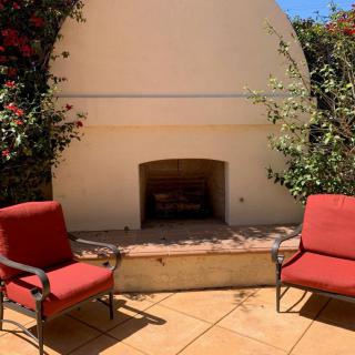 A sunny outdoor patio with a built-in fireplace and two red cushioned chairs, surrounded by lush greenery.