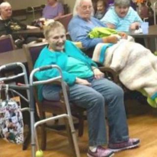 A group of elderly people gathered indoors, some using walkers and blankets.