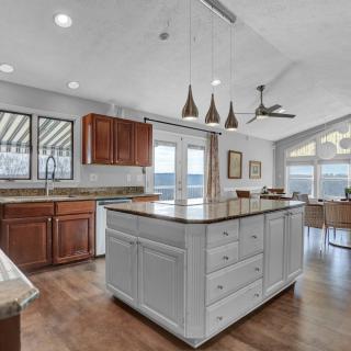 A modern kitchen with an island, featuring pendant lights and wooden cabinets, opens into a living area with large windows.