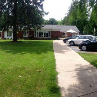A view of a brick house with a driveway, cars parked, and a well-maintained lawn surrounded by trees.