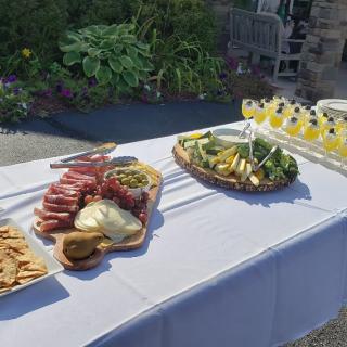 A sunny outdoor setting with a table displaying charcuterie, cheese, fruits, and drinks.