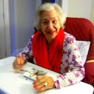 An elderly woman smiling warmly while seated at a table, wearing a red scarf and floral blouse.