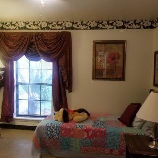 A cozy bedroom with a quilt-covered bed, an ornate window with curtains, and decorative elements including a lamp and paintings on the wall.