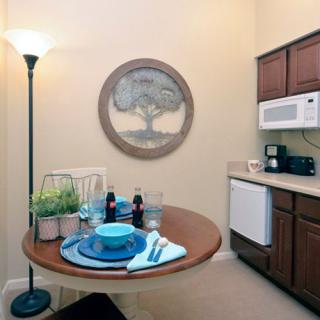 A cozy kitchen corner with a round table set for dining, featuring a lamp, wall artwork, and a compact kitchenette.