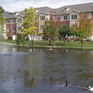 An apartment building next to a tranquil lake with ducks and a fountain, surrounded by trees.