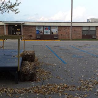 A quiet scene featuring a brick building with a parking lot scattered with autumn leaves and bales of hay.