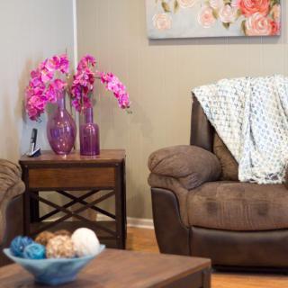 A cozy living room corner featuring a brown armchair with a decorative blanket and vibrant pink flowers in vases on a side table.
