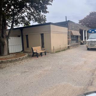 A view of a modest building with a bench in front and a parked vehicle nearby, set against a cloudy sky.