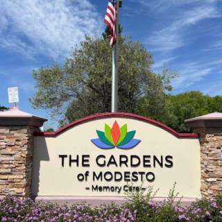 A sign for The Gardens of Modesto Memory Care facility, featuring landscaping and an American flag.