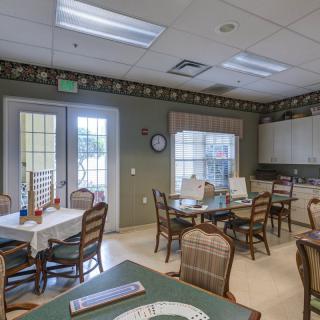 A cozy dining room with multiple tables and chairs, adjacent to a kitchen area, well-lit by natural light from the windows.
