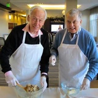 Two elderly men wearing aprons are engaged in a cooking activity together.