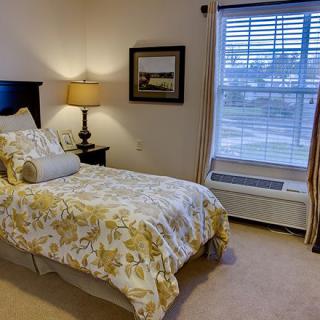 A cozy bedroom with a neatly made bed, a lamp on a side table, and natural light streaming through the window.